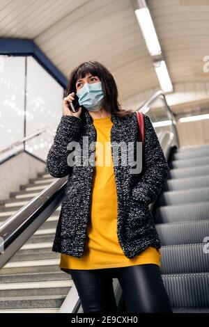 Photo de la femme heureuse portant un masque facial parlant au téléphone en se tenant dans les escaliers roulants. Banque D'Images