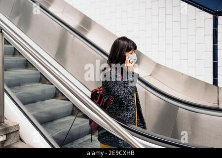 Photo de la femme heureuse portant un masque facial parlant au téléphone en se tenant dans les escaliers roulants. Banque D'Images