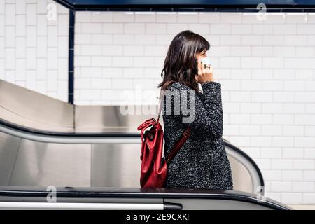 Photo de la femme heureuse portant un masque facial parlant au téléphone en se tenant dans les escaliers roulants. Banque D'Images