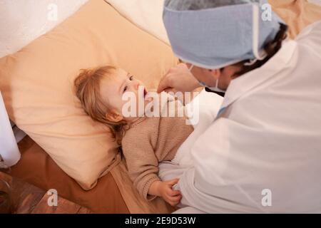 Le médecin vérifie la gorge d'une petite fille à la maison. Bébé malade, quarantaine à domicile, coronavirus, covid. Banque D'Images