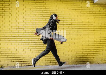 Moderne urbain jeune dreadlocks hippster homme courant avec son skate dans ses mains et musique d'écoute. Banque D'Images
