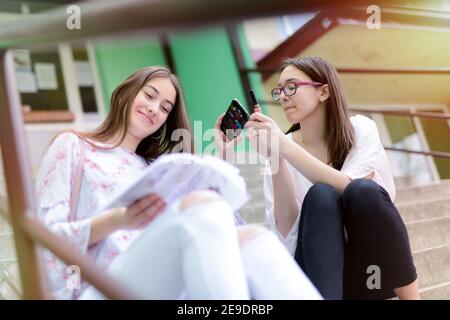 Deux jeunes filles d'école gaies assises sur les escaliers d'école et mettant le maquillage sur. L'un apprend alors que l'autre met son maquillage sur. Banque D'Images