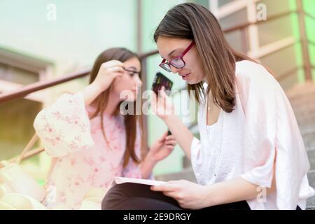 Deux jeunes filles d'école gaies assises sur les escaliers d'école et mettant le maquillage sur. L'un apprend alors que l'autre met son maquillage sur. Banque D'Images