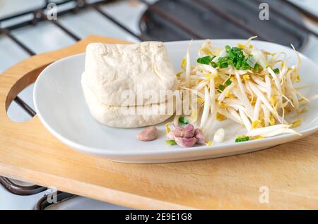 Pousses de haricots biologiques, tofu, garlics et échalotes en plaque blanche qui sur planche à découper en bois dans la cuisine, concept pour la cuisine végétarienne saine Banque D'Images