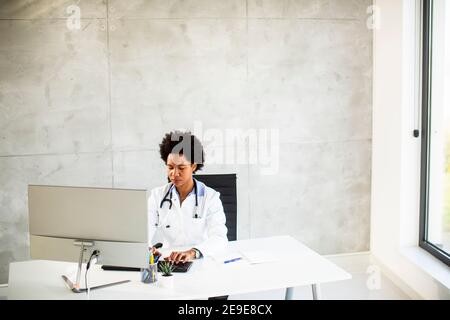 Femme afro-américaine médecin portant un manteau blanc avec stéthoscope assis derrière le bureau Banque D'Images