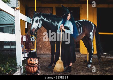 Une fille habillée comme une sorcière se tient par un corral sur une ferme à côté d'un cheval Banque D'Images