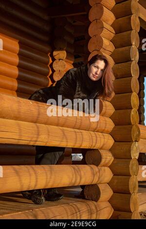 Une belle femme de 45 ans aux cheveux rouges pose sur la véranda d'une maison en rondins le jour d'hiver. Banque D'Images