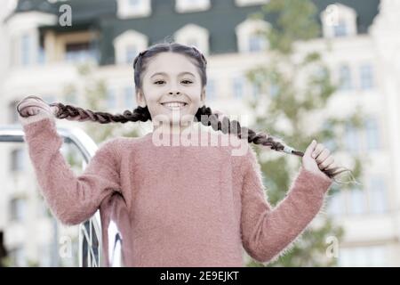 Avoir les cheveux en deux tresses. Adorable girl holding des tresses de cheveux sur l'arrière-plan urbain. Petit mignon fille avec de longs cheveux brune à l'extérieur. Peu de cheveux modèle avec look beauté. Banque D'Images