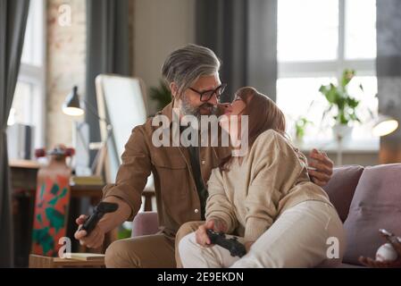 Couple d'âge mûr embrassant sur le canapé pendant leur jeu jeu vidéo dans le salon à la maison Banque D'Images