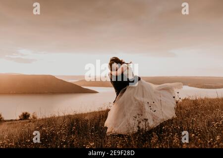 couple amoureux noces jeunes mariés en robe blanche et costume câlin embrassant le tourbillon sur l'herbe haute dans le champ d'été sur la montagne au-dessus de la rivière. coucher du soleil. Banque D'Images