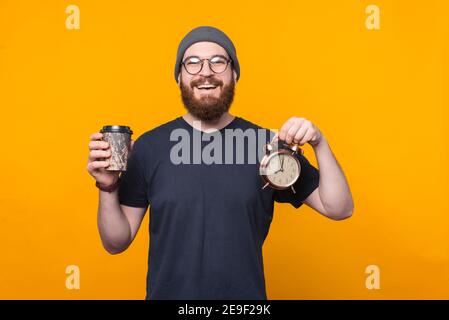 Portrait de l'homme hippster joyeux barbu tenant un réveil et une tasse de café, son temps de se détendre. Banque D'Images