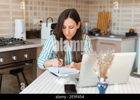 Photo d'une jeune femme prenant des notes dans un planificateur assis sur une table dans la cuisine. Banque D'Images