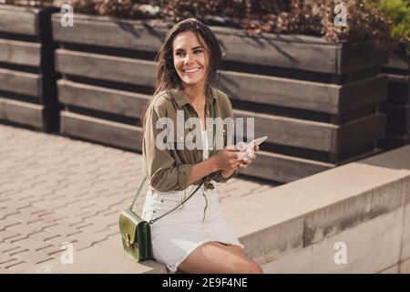 Photo portrait d'une fille gaie tenant le téléphone dans deux mains assis à l'extérieur du café, attendant que des amis viennent Banque D'Images