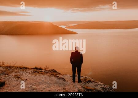 homme en costume habillé marié se tient sur la montagne tient ses mains dans les poches. orange vif coucher de soleil rayons de soleil à travers les nuages. vue panoramique de la falaise à Banque D'Images