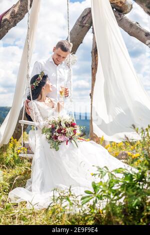 Un couple de jeunes mariés dans la nature buvant du champagne, une porte faite de matériaux naturels, un mariage dans la nature. Banque D'Images