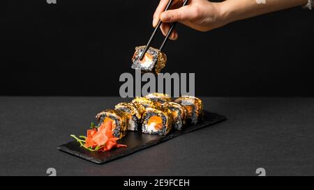 La main avec des baguettes noires contient un sushi. Rouleau de sushi personnalisé avec de l'encre de seiche, du saumon, du fromage à la crème, du poivre de citrouille, de l'anguille et des feuilles d'or comestibles avec Banque D'Images