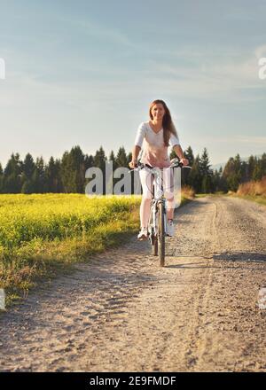 Une jeune femme fait du vélo sur une route poussiéreuse vers un appareil photo, le soleil de l'après-midi brille sur les champs et la forêt en arrière-plan Banque D'Images