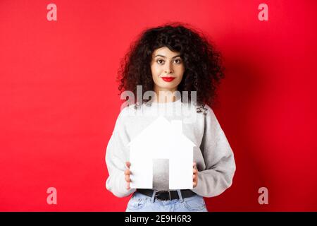 Immobilier. Jeune femme caucasienne vêtue de vêtements décontractés montrant une découpe de maison en papier, achetant des biens ou louant un appartement, debout sur fond rouge Banque D'Images