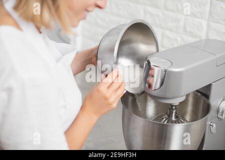Fouetter les blancs d'œufs avec du sucre dans le robot ménager. Mains de chef féminin professionnel ajoutant du sucre dans le bol de cuisine moderne en argent batteur sur socle Banque D'Images
