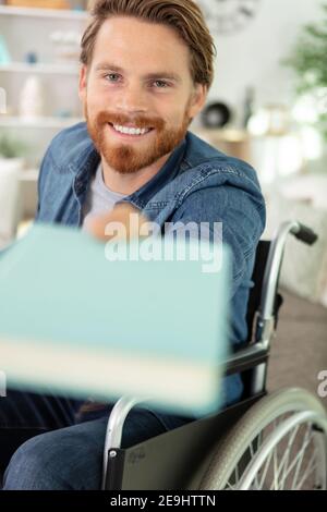 jeune homme joyeux en fauteuil roulant, passant un livre Banque D'Images