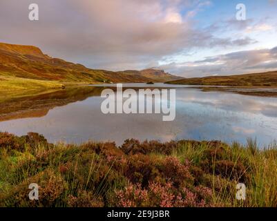 Île de Skye, Écosse : lumière du lever du soleil et nuages se reflétant dans le Loch Fada avec des bruyères qui fleurissent sur le rivage et l'emblématique Old Man of Storr dans le Th Banque D'Images