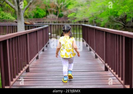 Une vue arrière d'une petite fille asiatique qui marche seule le long d'un chemin dans un parc avec rail en bois des deux côtés. Banque D'Images