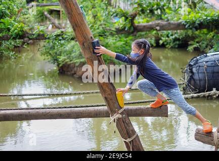 Une jolie jeune fille asiatique essayant de traverser un petit canal à travers une structure en bois avec des trous d'ouverture, en étant courageuse mais prudente pour atteindre sa destination. Banque D'Images