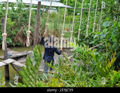 La vue arrière d'une jolie jeune fille asiatique essayant de traverser un petit canal en se équilibrant une bûche avec une corde sur sa tête pour l'aider Banque D'Images
