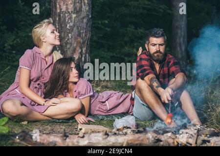 Amis campant en forêt. Filles et garçon espiègles avec un regard sérieux assis près d'un feu de camp et de saucisses volantes. Banque D'Images