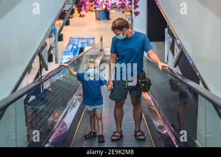 Vue de face garçon, père monter escalator dans super marché porter masque de protection du visage, COVID coronavirus de quarantaine de confinement pandémique. Papa tient la main de l'enfant Banque D'Images