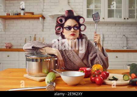 Drôle de femme de maison en colère dans les rouleaux de cheveux assis à la table de cuisine et en attente de mari Banque D'Images