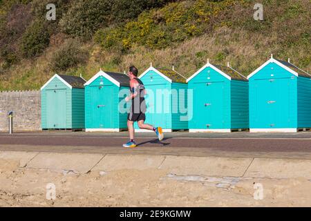 Bournemouth, Dorset, Royaume-Uni. 5 février 2021. Météo au Royaume-Uni : le soleil chaud s'est envengé avec des douches sur les plages de Bournemouth tandis que les gens se dirigent vers le bord de mer pour leur exercice quotidien pendant le Lockdown 3 de l'épidémie pandémique du coronavirus Covid 19 Covid19 du virus Corona. Des pistes de jogging Man JOGS longent la promenade et longent des cabanes de plage. Crédit : Carolyn Jenkins/Alay Live News Banque D'Images