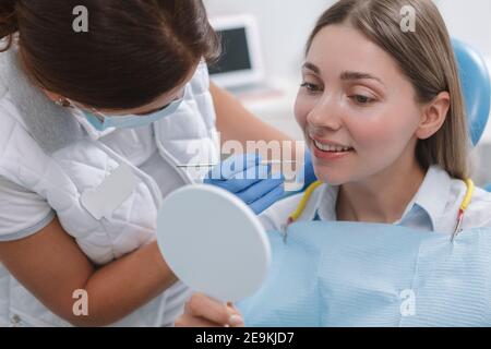 Belle jeune femme regardant dans le miroir pendant le traitement dentaire par dentiste Banque D'Images
