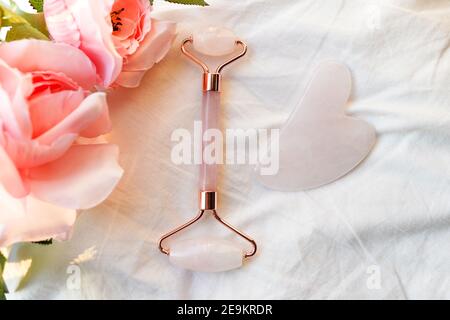 rouleau de visage rose et masseur de gua sha en pierre néphrite de jade naturelle et fleurs roses sur fond blanc. Concept de massage du visage. Banque D'Images