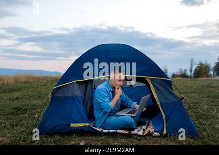 Homme travaillant sur un ordinateur portable dans une tente dans la nature. Jeune indépendant assis dans le camp. Détente en montagne. Travail à distance, activités en plein air en été. Joyeux homme Banque D'Images