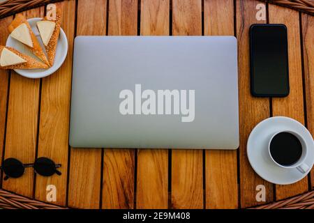 Un espace de travail sur une table en bois naturel, avec un décor minimaliste. Ordinateur portable fermé, smartphone, tasse à café, cookies et lunettes élégantes Banque D'Images