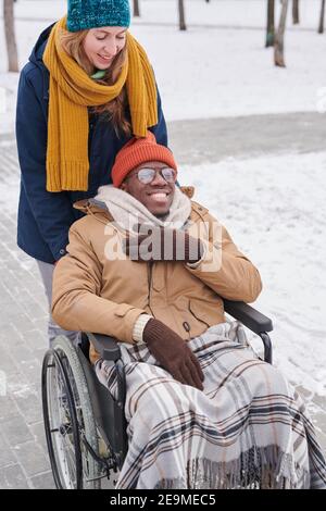 Heureux Africain assis en fauteuil roulant et en appréciant sa marche avec un ami sur l'air frais en hiver Banque D'Images