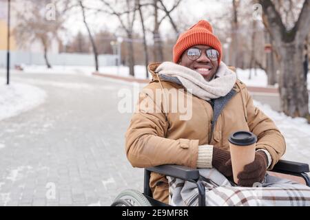 Portrait d'un homme africain handicapé en fauteuil roulant souriant à la caméra tout en buvant du café chaud dans le parc Banque D'Images