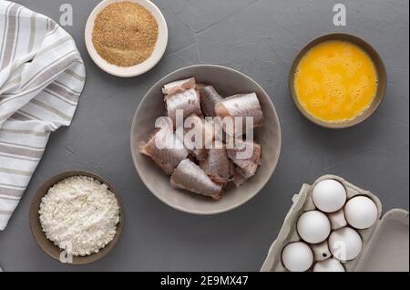 Dans un bol, des morceaux de merlu et des produits pour la cuisine frite, des œufs, de la farine et de la chapelure ont été présentés avec une serviette de cuisine sur fond gris Banque D'Images