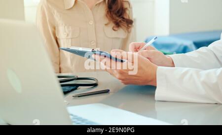 Gros plan mains de Male Doctor remplit une carte de patient, jeune femme patiente assise près de l'arrière-plan. Parler avec un médecin à la consultation Banque D'Images