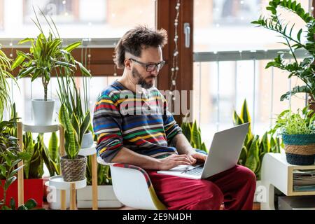 Jeune homme travaillant à la maison avec un ordinateur portable entouré de plantes de maison Banque D'Images