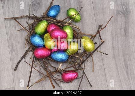 Oeufs de pâques multicolores enveloppés de feuilles en rose, vert, bleu et jaune dans un nid naturel fait de bâtons et de brindilles, sur un fond en bois gris. Banque D'Images