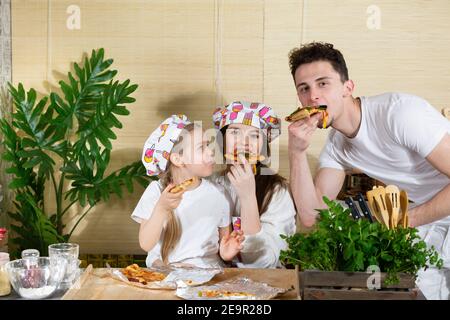 Afin d'être plus proche de maman, la fille s'est assise sur ses genoux et le père a embrassé sa mère et ensemble ils ont commencé à manger leur pizza fraîche cuite. La joie de maman, papa et fille se réunit dans la cuisine. Banque D'Images