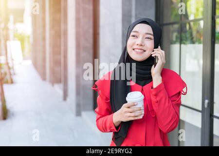 Femme d'affaires musulmane asiatique heureuse d'utiliser le téléphone intelligent pour appeler le contact client en marchant dans la rue avec une tasse de café Banque D'Images