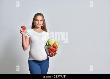 Femme blonde enceinte tenant des légumes frais dans un bol et tomate dans sa main Banque D'Images