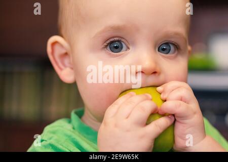 Visage de bébé de race blanche avec pomme. Gros plan. Première nourriture pour bébé. L'enfant se salit le visage en mangeant des fruits. Pomme verte fraîche dans la main de bébé. Banque D'Images