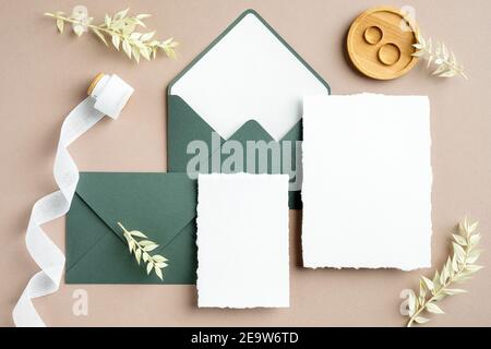 Élégante papeterie de mariage. Modèles de cartes d'invitation de mariage, enveloppes vertes, ruban en soie, anneaux dorés sur fond beige pastel. Pose à plat, t Banque D'Images