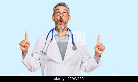 Homme mi-âge à cheveux gris portant l'uniforme de médecin et le stéthoscope stupéfait et surpris regardant vers le haut et pointant avec les doigts et les bras relevés. Banque D'Images