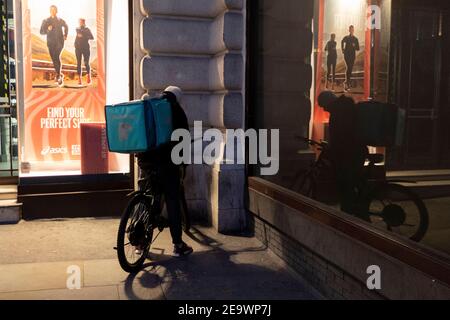 Un pilote de livraison de Deliveroo trouve un coin sombre d'un Piccadilly Circus lumineux pour vérifier son téléphone lors du troisième verrouillage de la pandémie de coronavirus, le 5 février 2021, à Londres, en Angleterre. Banque D'Images