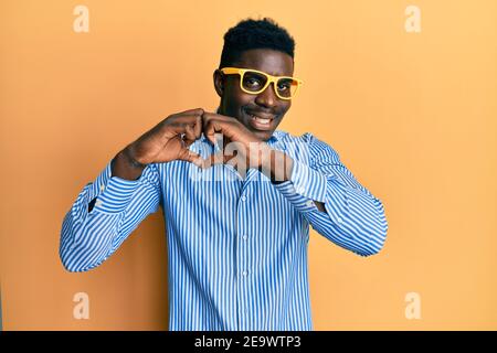 Beau noir homme portant des lunettes jaunes souriant dans l'amour faisant coeur symbole de forme avec les mains. Concept romantique. Banque D'Images